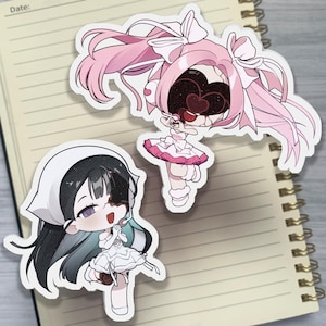 May include: Two die-cut vinyl stickers featuring anime-style characters. One character has long pink hair with white bows and a dark, heart-shaped void for a face. The other character has black hair, a white head covering, and a dark patch over one eye.