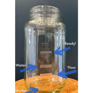 Puede incluir: Un tarro Mason de vidrio transparente con etiquetas para agua, harina y masa madre. El tarro tiene una tapa de rosca y la palabra "Ready!" está escrita en el lateral. El tarro está sobre una superficie marrón.