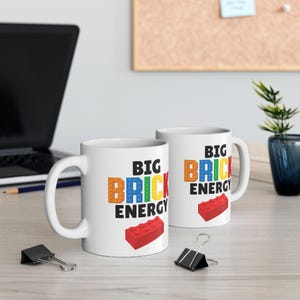 Peut inclure: Deux tasses en céramique blanche avec le texte BIG BRICK ENERGY écrit en lettres noires et colorées style briques. Sous le texte, une brique de construction rouge est illustrée. Les tasses ont une anse standard et une finition lisse.