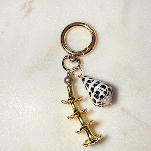 May include: A gold-toned metal keychain featuring a circular ring, a small grey bead, a vertical gold-toned branch-like charm with three tiers of curved protrusions, and a single cone-shaped seashell with a black and white checkered pattern.