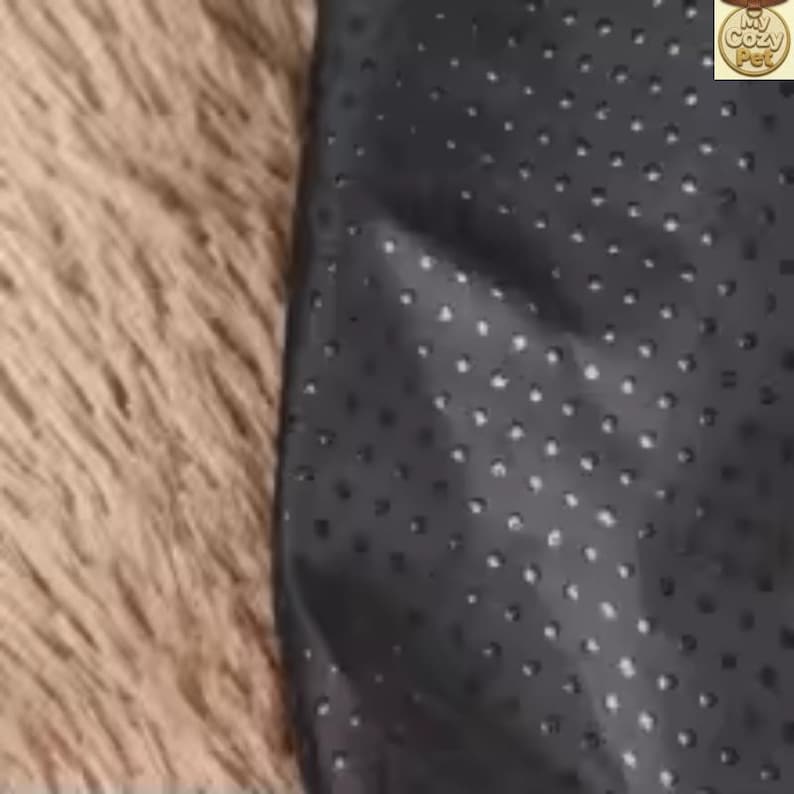 May include: A close-up of a dark grey pet bed with a textured, dotted surface. The bed is next to a fluffy, light brown surface. The image is well-lit, highlighting the contrast between the textures and colours.