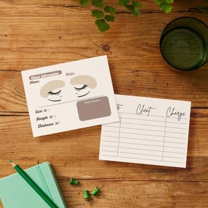 May include: Two cream-colored client information cards and a charge sheet rest on a wooden surface. The client card features a stylized eye design and prompts for curl, length, and thickness. A green pencil and notebook are also present.