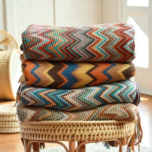 May include: Five folded blankets with a chevron design in shades of red, teal, brown, and yellow. The blankets are stacked on a round wicker stool. The blankets have a soft appearance. The stool is made of woven wicker.