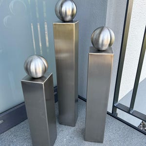 Könnte beinhalten: Drei moderne Außenskulpturen. Jede Skulptur besteht aus einer rechteckigen Säule aus gebürstetem Metall, die von einer kugelförmigen, gerippten Metallkugel gekrönt wird. Die Skulpturen sind in einer Gruppe angeordnet, unterschiedlich hoch und vor einem neutralen Hintergrund platziert.