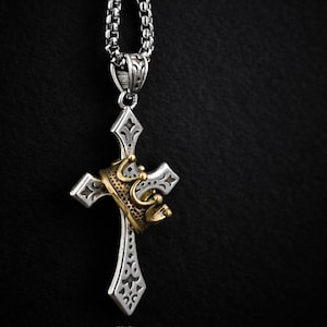 May include: Silver-toned cross pendant with a gold-colored crown accent. The cross features intricate detailing and hangs from a matching chain. The pendant is set against a dark background, highlighting the contrast of the metals.