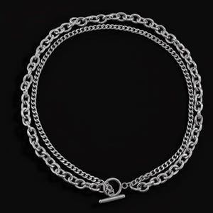 May include: A silver-toned double-layered chain necklace featuring a thick oval link chain and a thinner curb chain. The necklace is secured with a circular toggle clasp at the bottom, which includes a horizontal bar that passes through the ring.