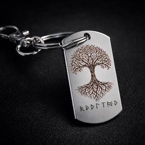 May include: A silver-colored dog tag keychain with a detailed tree design etched in brown. The tree has intricate branches and roots. Below the tree is the word "ROOTED" in a runic font. The keychain has a clasp.