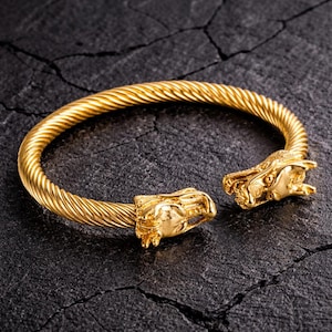 May include: A gold-colored cuff bracelet with a twisted rope design. The bracelet features dragon head terminals. The bracelet is open, revealing the intricate details of the dragon heads and the twisted metal.