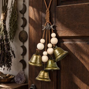May include: A decorative hanging with brass bells, wooden beads, and a silver charm with a crescent moon and star design. The bells are suspended from a braided cord, creating a rustic and mystical aesthetic. The background is a wooden door.