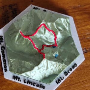 May include: A 3D-printed topographical map of a mountain range, featuring green terrain with a red trail and blue lakes. The map is set on a white hexagonal base with the names "Mt. Cameron", "Mt. Lincoln", and "Mt. Bross" in black text.