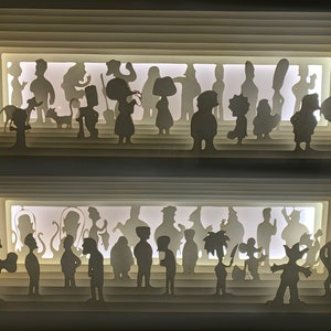 May include: A white papercraft art piece with a layered design featuring silhouettes of people. The piece is illuminated from behind, creating a glowing effect. The silhouettes are arranged in two rows, with the top row featuring a group of people standing together and the bottom row featuring a group of people standing in a line.