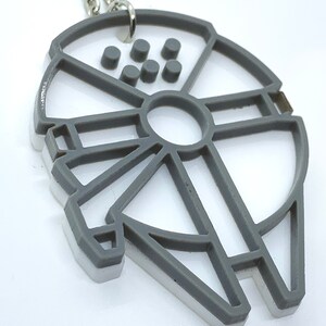 May include: A gray and white plastic keychain shaped like the Millennium Falcon from Star Wars. The keychain has a silver loop for attaching to keys.