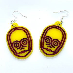 May include: A pair of yellow earrings with a brown outline of a robot face. The earrings are made of acrylic and have silver hooks.
