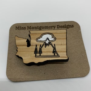 May include: Wooden cutout of the state of Washington with a mountain, cloud, and trees inside. The cutout is on a brown background with the text "Miss Montgomery Designs" at the top.