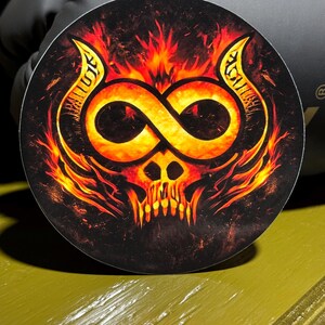 May include: Round sticker featuring a fiery skull design. The skull has an infinity symbol in place of the eyes and is surrounded by flames in shades of orange and yellow. The sticker has a black background.
