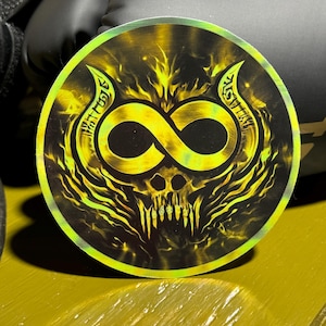 May include: A round sticker with a yellow and black design featuring an infinity symbol, a skull, and horns. The sticker has a holographic effect, with a green and yellow iridescent sheen. The background is black.