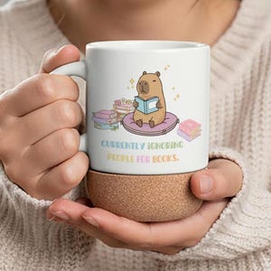 Könnte beinhalten: Eine weiße Keramik-Tasse mit schwarzem Henkel und dem Text "But First, Coffee" in schwarzer, handschriftlicher Schrift. Die Tasse steht auf einer weißen Oberfläche.