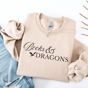 Könnte beinhalten: Beiges Sweatshirt mit dem schwarzen Schriftzug "Books & Dragons" und einer Drachen-Grafik. Eine silberne Halskette mit kleinem Anhänger liegt auf dem Sweatshirt, das auf einer blauen Jeans gefaltet ist.