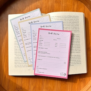 Könnte beinhalten: Vier rechteckige Buchbewertungskarten in Hellgrün, Gelb, Blau und Pink mit einem geometrischen Randmuster. Jede Karte enthält den Text Book Review, ein Herzsymbol sowie vorgedruckte Linien für Titel, Autor, Bewertung und Leseanmerkungen.