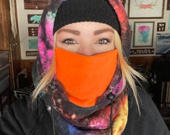 May include: A woman wearing a black hooded sweatshirt with a colorful galaxy print fleece neck gaiter and an orange fleece face mask. The text "ROXY" is visible on the sweatshirt.