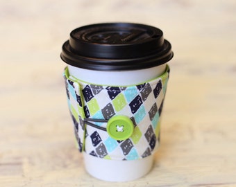 May include: A white coffee cup with a black lid and a green, blue, and gray patterned fabric sleeve with a green button.