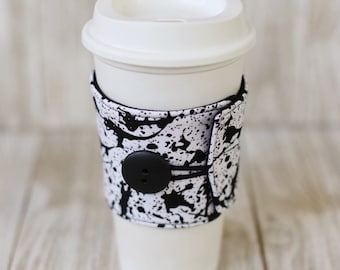 May include: A white coffee cup with a black and white splatter pattern fabric sleeve. The sleeve has a black button and elastic band.