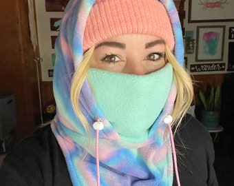 May include: A tie-dye fleece neck gaiter with pink drawstrings and white toggles. The gaiter is worn over a pink knit beanie.
