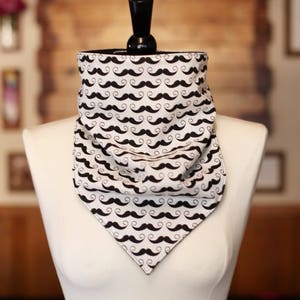 May include: A white and black mustache patterned neck gaiter displayed on a mannequin. The neck gaiter is made of a soft fabric and features a repeating pattern of black mustaches on a white background. The neck gaiter is designed to be worn around the neck.