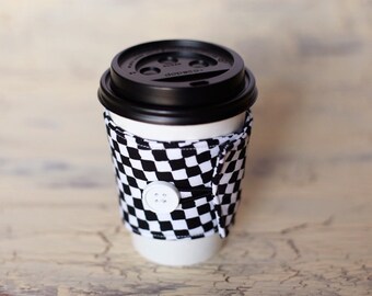 May include: A black and white checkered coffee cup cozy with a white button closure. The cozy is on a white paper cup with a black lid.