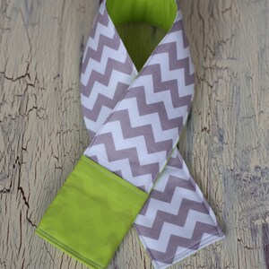 May include: A gray and white chevron patterned fabric neck warmer with a lime green lining. The neck warmer is folded in half and the lining is visible on the outside.