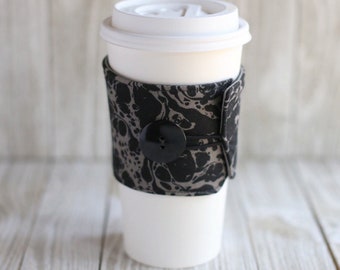 May include: A white coffee cup with a black and gray fabric sleeve with a black button. The sleeve has a marbled pattern.