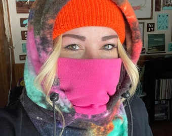 May include: A woman wearing a black hooded fleece neck gaiter with a galaxy print. The gaiter has a pink fleece lining and a black drawstring with black toggles. The woman is also wearing an orange knit beanie.