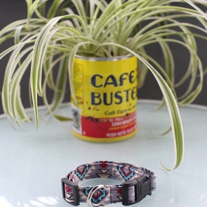 May include: A gray and blue patterned dog collar with a silver buckle. The collar is on a white surface with a plant in a yellow can that says "Cafe Buste" in the background.