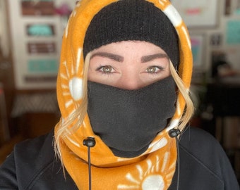 May include: A yellow fleece neck gaiter with a white sun design. The gaiter has a black face mask attached and black drawstrings with black toggles. The gaiter is worn over a black beanie and a black sweatshirt with the word "ROXY" printed on the left side of the chest.