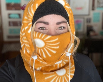 May include: A yellow fleece neck gaiter with a white sunburst pattern. The gaiter has a white drawstring with white toggles. The gaiter is worn by a person with a black beanie and a black sweatshirt with the word "ROXY" printed on the left side.