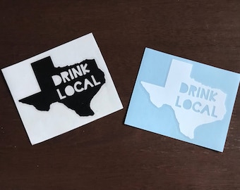 May include: Two square stickers with a Texas outline and the words "DRINK LOCAL". One sticker is black with white text, the other is white with light blue background and text. The stickers are on a brown surface.