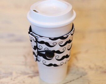 May include: A white coffee cup with a black and white patterned fabric sleeve. The sleeve has a black button and elastic band. The pattern on the sleeve is a repeating design of mustaches and swirls.