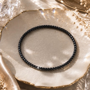 May include: A thin, circular bracelet composed of small, uniform, square-shaped black beads with a polished, reflective finish. The beads are strung together in a continuous loop, creating a sleek and minimalist design that sits in a circular form.