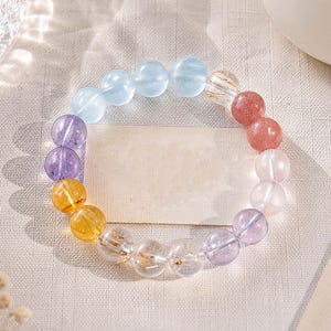 Può includere: Un braccialetto di perline con una disposizione circolare di sfere di pietre preziose lisce e levigate in un gradiente di colori che include azzurro, viola, giallo, trasparente con inclusioni dorate, rosa e lavanda, su un cordino elastico.