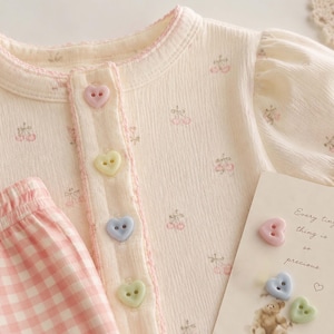 Puede incluir: Una prenda superior de color crema con estampado de pequeñas cerezas, cuello redondo con borde rosa y cuatro botones pastel en forma de corazón. Junto a ella, una prenda de cuadros vichy rosa y blanco y una tarjeta con el texto "Every tiny thing is so precious."
