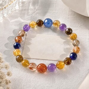 May include: A beaded bracelet featuring a variety of polished, spherical gemstones in shades of purple, yellow, brown, orange, and blue. The beads are arranged in a circular pattern, showcasing natural inclusions and translucent textures throughout.