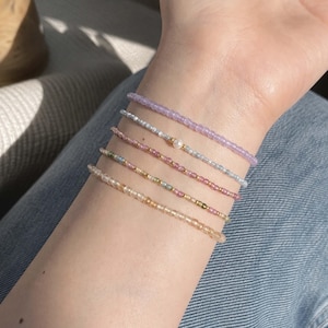 Può includere: Un set di cinque delicati braccialetti di perline indossati al polso. I braccialetti presentano piccole perline traslucide nei toni del lavanda, azzurro, rosa, multicolore e ambra. Un braccialetto azzurro include una singola perla bianca.
