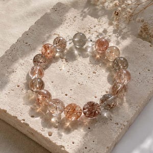 Puede incluir: Una pulsera circular compuesta por cuentas esféricas translúcidas con inclusiones internas en forma de aguja en tonos cobre, marrón y negro. Las cuentas están dispuestas en un bucle continuo, intercaladas con una cuenta redonda, lisa y clara.