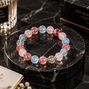 Può includere: Un braccialetto di perline con pietre rotonde levigate che alternano tonalità azzurre, rosa tenue e rosso traslucido con inclusioni maculate. Le perline sono infilate con piccoli distanziatori in metallo dorato tra ogni singola pietra.