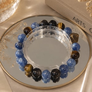 Könnte beinhalten: Ein kreisförmiges Perlenarmband mit abwechselnd polierten, kugelförmigen Perlen in den Farben Tiefblau, Goldbraun und Schwarz. Das Armband ist in einer durchgehenden Schleife angeordnet und zeigt die glatte, reflektierende Oberfläche jeder einzelnen Perle.