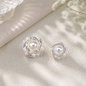May include: Two circular stud earrings featuring a central white faux pearl surrounded by a twisted silver-tone metal frame and smaller white faux pearls. The design is symmetrical with a textured metallic finish and a polished pearl center.