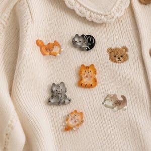 May include: A cream-colored knit cardigan featuring a scalloped collar and brown buttons, adorned with several small, translucent, cat-shaped buttons in orange, gray, and brown, alongside a single embroidered brown bear face patch.