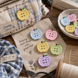 May include: A collection of apple-shaped buttons in pastel gingham patterns including yellow, blue, pink, green, and purple. Each button features four holes and a small green stem. The buttons are displayed alongside a fabric pouch with the text Sewing small things with great love, a blue gingham pouch labeled Cuddly Natural life, and a bowl of additional buttons.