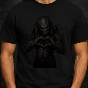 May include: A black short-sleeved t-shirt featuring a dark, monochromatic graphic print of the Predator character. The creature is depicted in a textured, armored suit with its signature dreadlocks, forming a heart shape with its hands.
