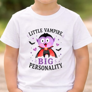 May include: A white short-sleeved t-shirt featuring a graphic of a purple vampire character with a red-lined black cape. The text reads LITTLE VAMPIRE, BIG PERSONALITY, surrounded by small black bats, purple hearts, and star-shaped accents.